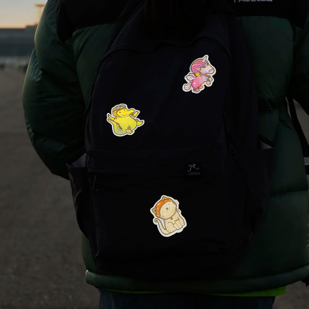Reflektierende Aufkleber Textil Stoff Einhorn „Juno“,  Krokodil „Freddy“ und Katze „Miez“  aus der Produktserie „Reflective Friends“  auf einem schwarzen Rucksack der von einem Kind getragen wird. Von REFLECTIVE Berlin.