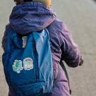 Reflektierende Aufkleber Stoff Textil Oktopus „Otto“ , Dinosaurier T-Rex „Lino“ und Eule „Olli“ aus der Produktserie „Reflective Friends“  die auf einen blauen Rucksack geklebt sind der von einem Kind auf einem Roller getragen wird. Von REFLECTIVE Berlin.