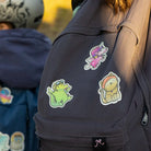 Reflektierende Aufkleber Textil Stoff Krokodil „Freddy“, Einhorn „Juno“ und Katze „Miez“ aus der Produktserie „Reflective Friends“  die auf einen schwarzen Rucksack geklebt sind der von einem Kind getragen wird. Ein weiteres Kind ist unscharf im Hintergrund. Von REFLECTIVE Berlin.