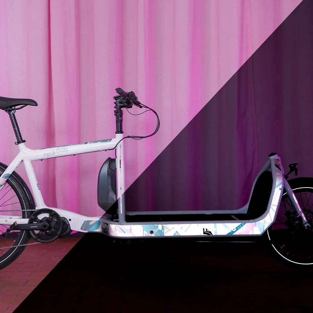 Reflektierende Aufkleber Folie XXL „Flamingo Funk“ individuell zugeschnitten und auf dem Rahmen von einen Bullitt  Cargo Fahrrad in Farbe "Milk Plus“  verklebt sind. Das Bild ist zweigeteilt und zeigt auf der linken Seite das Fahrrad und Sticker ohne Reflexion, auf der rechten Seite sind die zugeschnittenen Sticker mit Reflexion im Dunkeln dargestellt. Arrangement von REFLECTIVE Berlin.