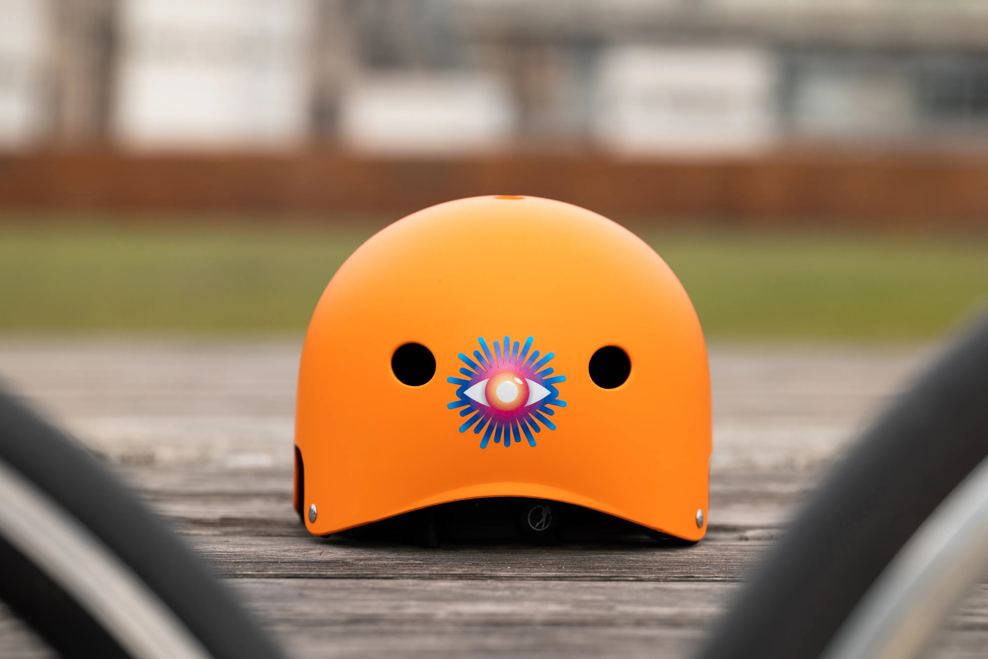 Reflektierender Aufkleber „Auge“ in Farbvariante „Regenbogen“ auf orangem Fahrradhelm Rückansicht vor unscharfen Hintergrund Detailansicht. Von REFLECTIVE Berlin.