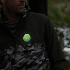 grüner reflektierender Button Anstecker „Berge“ auf einer Regenjacke in oliven Tarnmuster, die von einer Person getragen wird. Arrangement bei Dämmerung mit Reflexion des Buttons von REFLECTIVE Berlin.