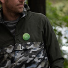 grüner reflektierender Button Anstecker „Berge“ auf einer Regenjacke in oliven Tarnmuster, die von einer Person getragen wird. Arrangement bei Tageslicht von REFLECTIVE Berlin.