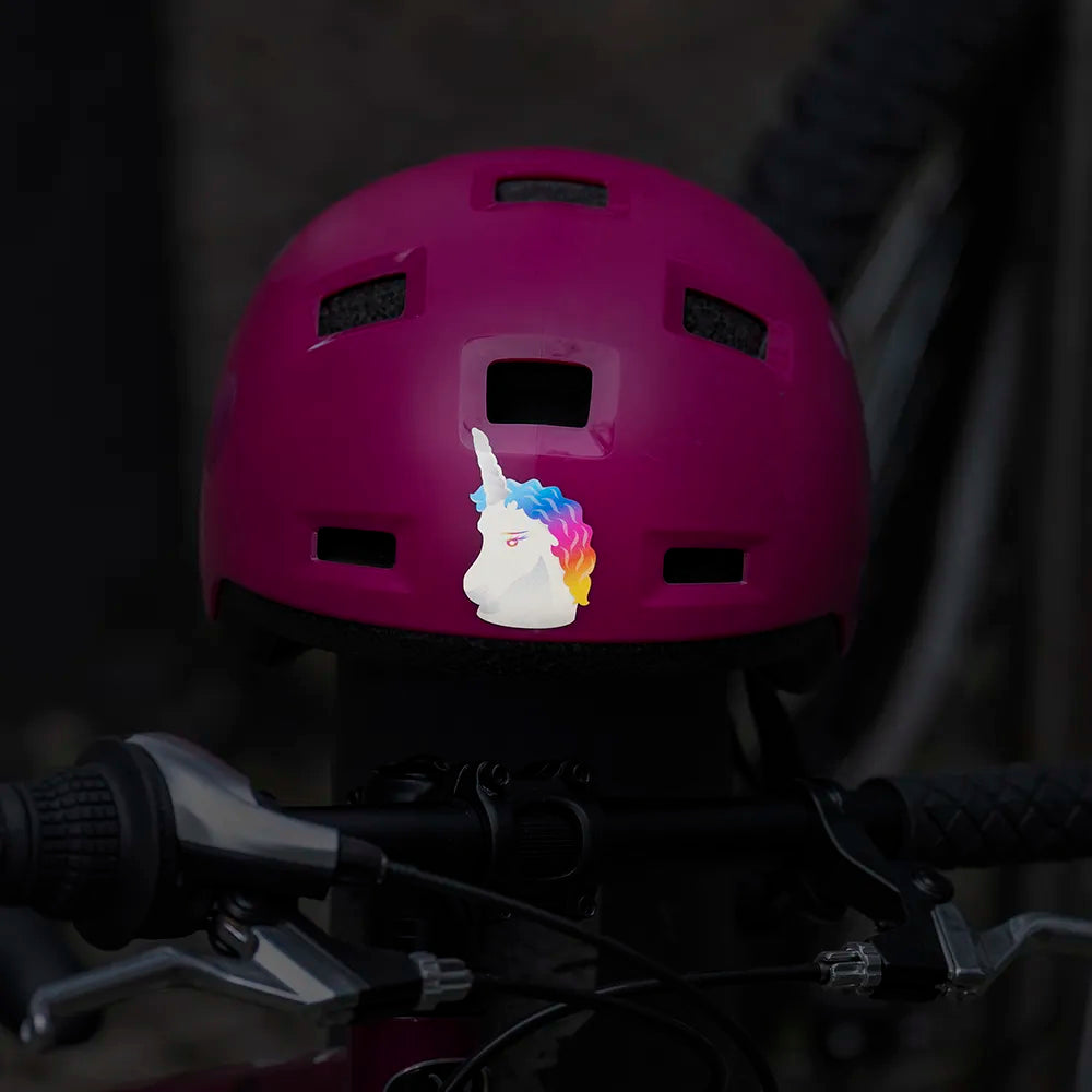Reflektierender Aufkleber „Einhorn“ in Farbvariante „Regenbogen“ auf pinken Fahrradhelm vor dunklem Hintergrund. Von REFLECTIVE Berlin.