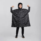 reflektierender Fahrrad Regenponcho in Farbvariante „Doodle“ getragen von einer Person, die frontal in die Kamera schaut und die Arme hebt, vor weißem Hintergrund. Kollaboration von Bomance und REFLECTIVE Berlin.