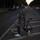 stark reflektierender Fahrrad Regenponcho in Farbvariante „Doodle“ getragen von einer Person im Profil, die einen Fahrradhelm trägt und mit einem Fahrrad über eine Straße geht. Kollaboration von Bomance und REFLECTIVE Berlin.