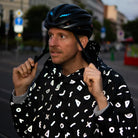 stark reflektierender Fahrrad Regenponcho in Farbvariante „Doodle“ getragen von einer Person, die einen Fahrradhelm trägt und über eineStraße geht. Die Doodle Elemente reflektieren stark. Kollaboration von Bomance und REFLECTIVE Berlin.