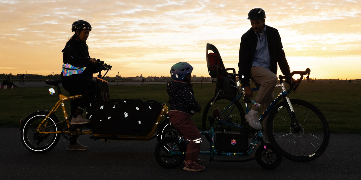 Familie auf dem Weg vom Spielplatz nach Hause im Sonnenuntergang. Mutter fährt mit Lastenrad, Vater mit Gravelbike, Tochter mit Laufrad. Alle haben reflektierende Designs an den Rädern und an der Kleidung um im Dunklen gut gesehen zu werden.