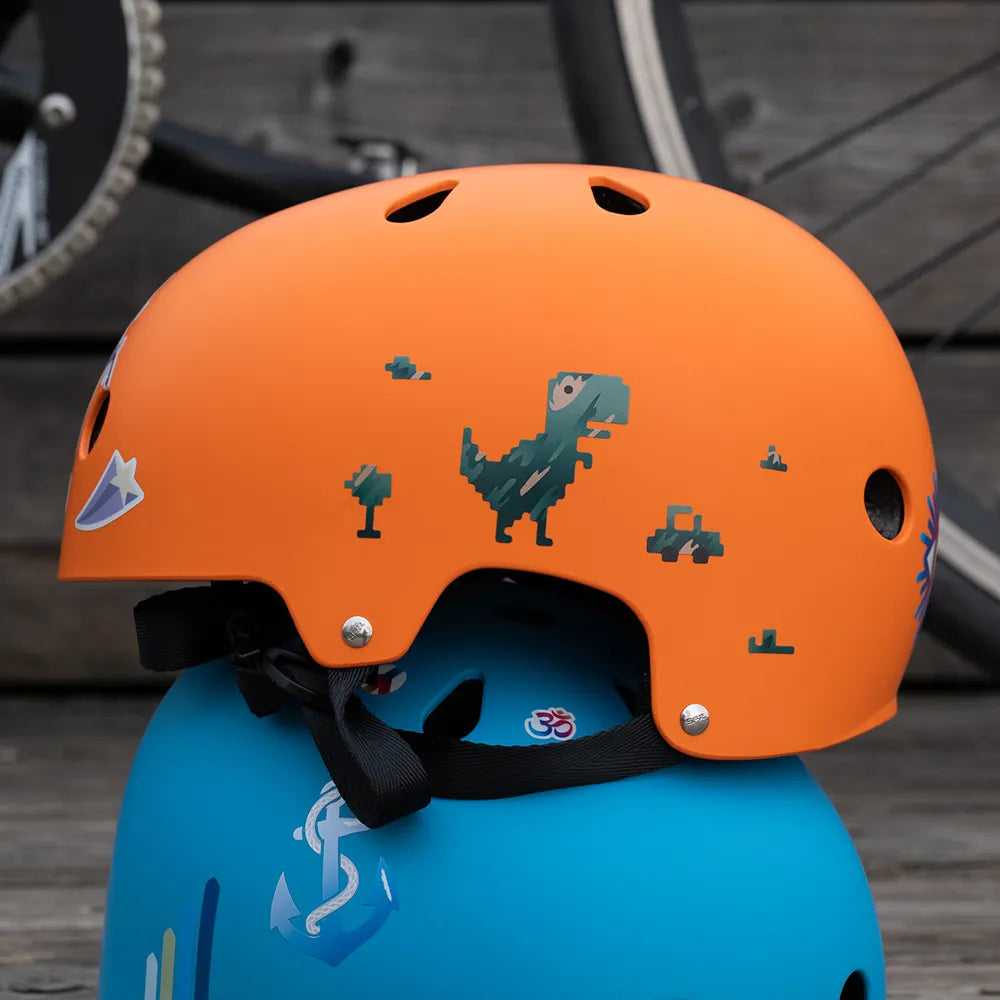 Reflektierender Aufkleber „T-Rex“ in Farbvariante „camo“ auf orangem Fahrradhelm der auf einem blauen Fahrradhelm liegt. von REFLECTIVE Berlin.