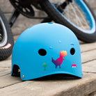 Reflektierender Aufkleber „T-Rex“ in Farbvariante „Regenbogen“ auf blauem Fahrradhelm vor unscharfen Hintergrund. Von REFLECTIVE Berlin.