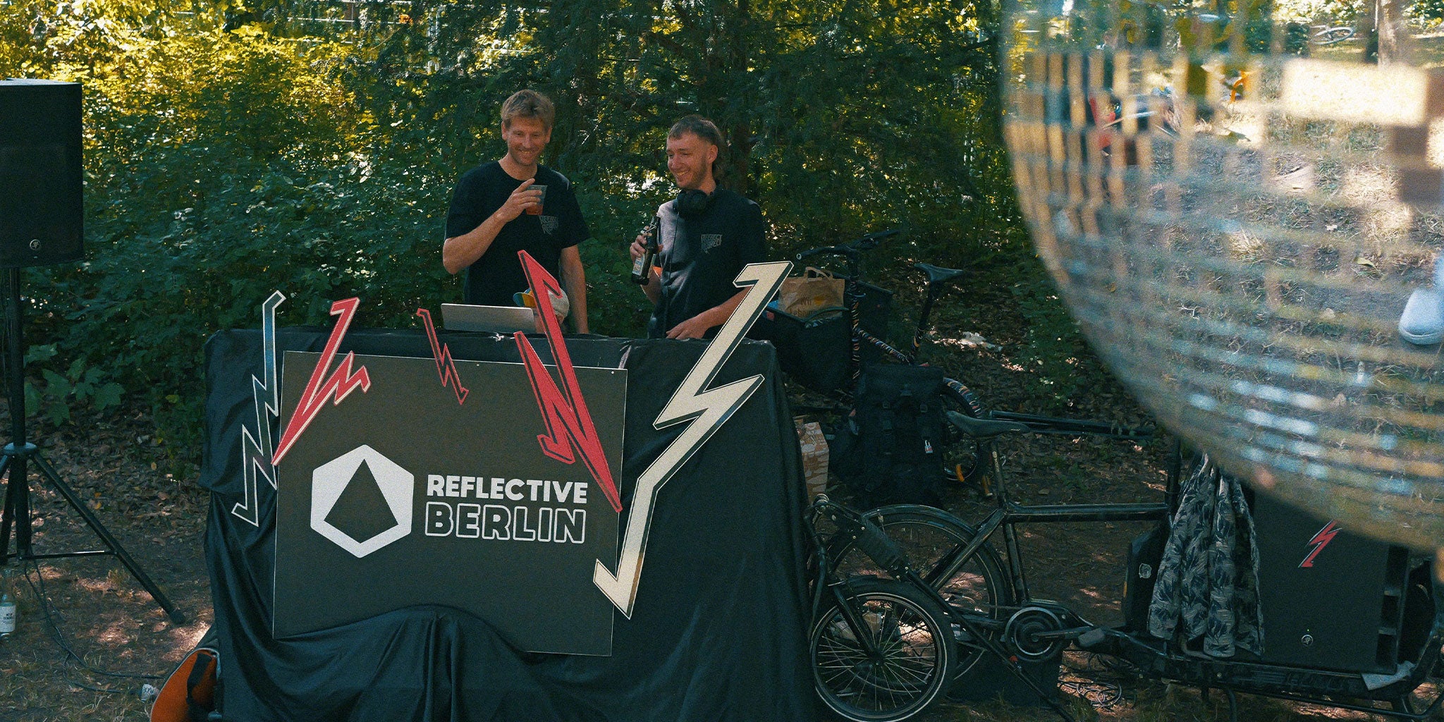 2 DJs stehen im Park hinterm DJ Pult und lächeln
