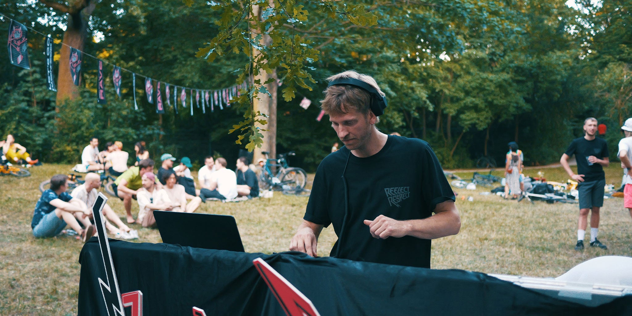 DJ im Park guckt auf seinen Laptop. Im Hintergrund sitzen Menschen auf der Wiese