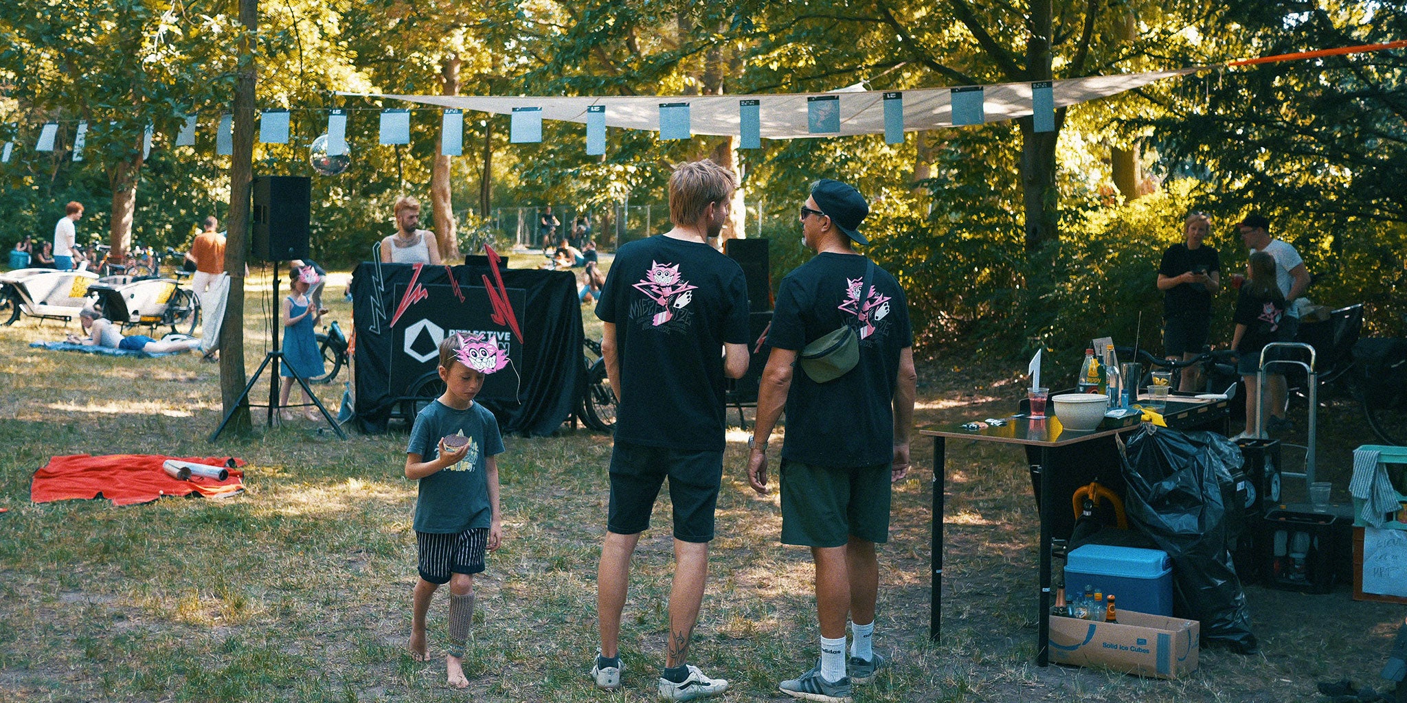 2 Männer von hinten im Park, die sich unterhalten. Links von ihnen ein Kind mit Reiswaffel und Katzenmaske. Im Hintergrund ein DJ am Pult