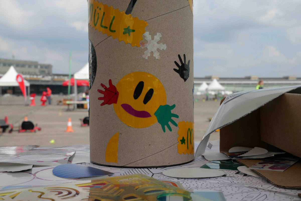 Eine mit Stickern und einem Smiley beklebte Papphülse steht auf einem Basteltisch und im Hintergrund erkennt man Pavillons auf dem Gelände des Tempelhofer Flughafens