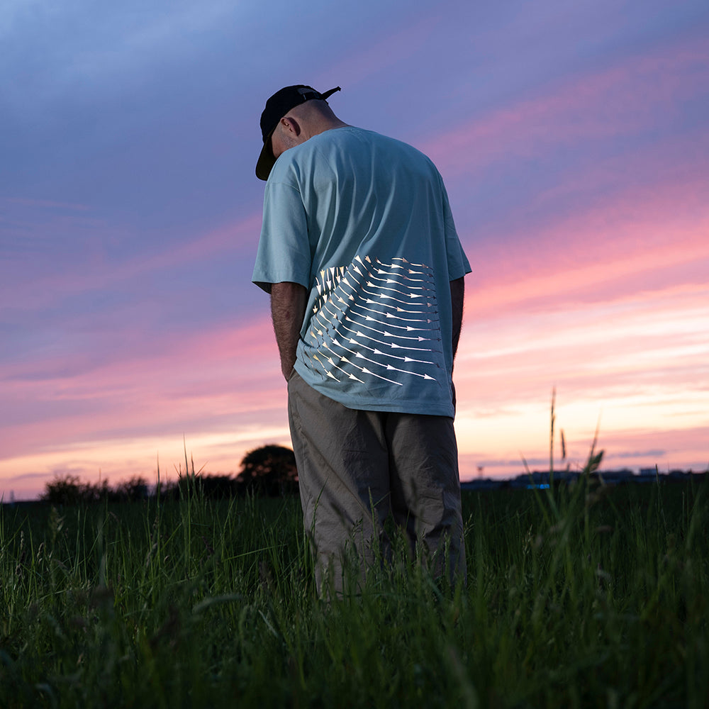 Mann im Feld trägt ein beige Hose und blaues Shirt mit stark reflektierendem Muster, schaut hinab