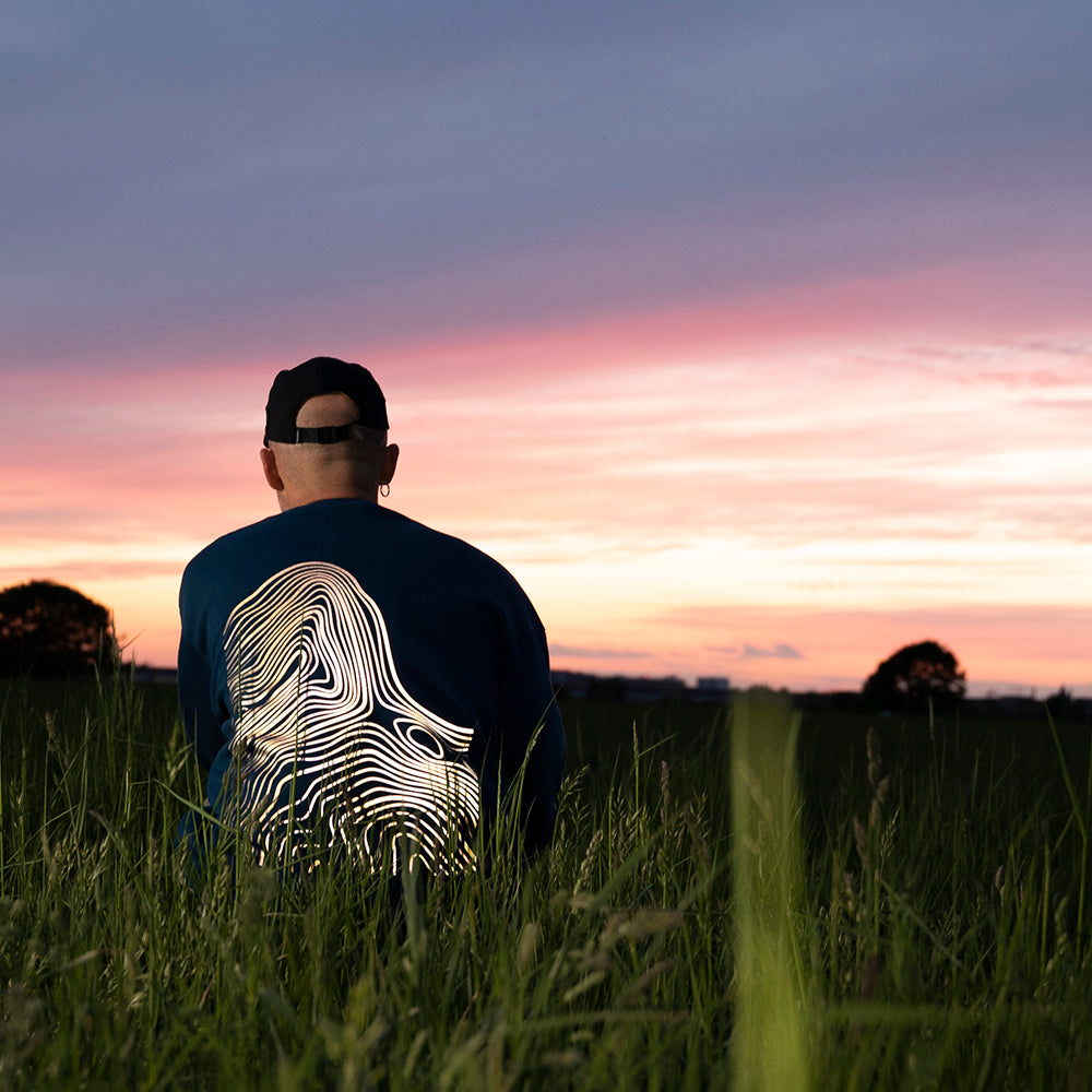 Kerl schaut in den Sonnenuntergang und hockt im Grad, trägt Pulli mit großem reflektierendes Muster