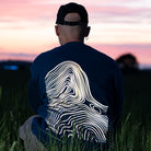 Mann mit Cap und reflektierendem Pullover hockt im Gras, Rückenansicht
