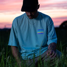 Typ mit Cap kniet im Gras, schaut runter, sein mintfarbenes T-Shirt hat bunte Streifen auf der Brust