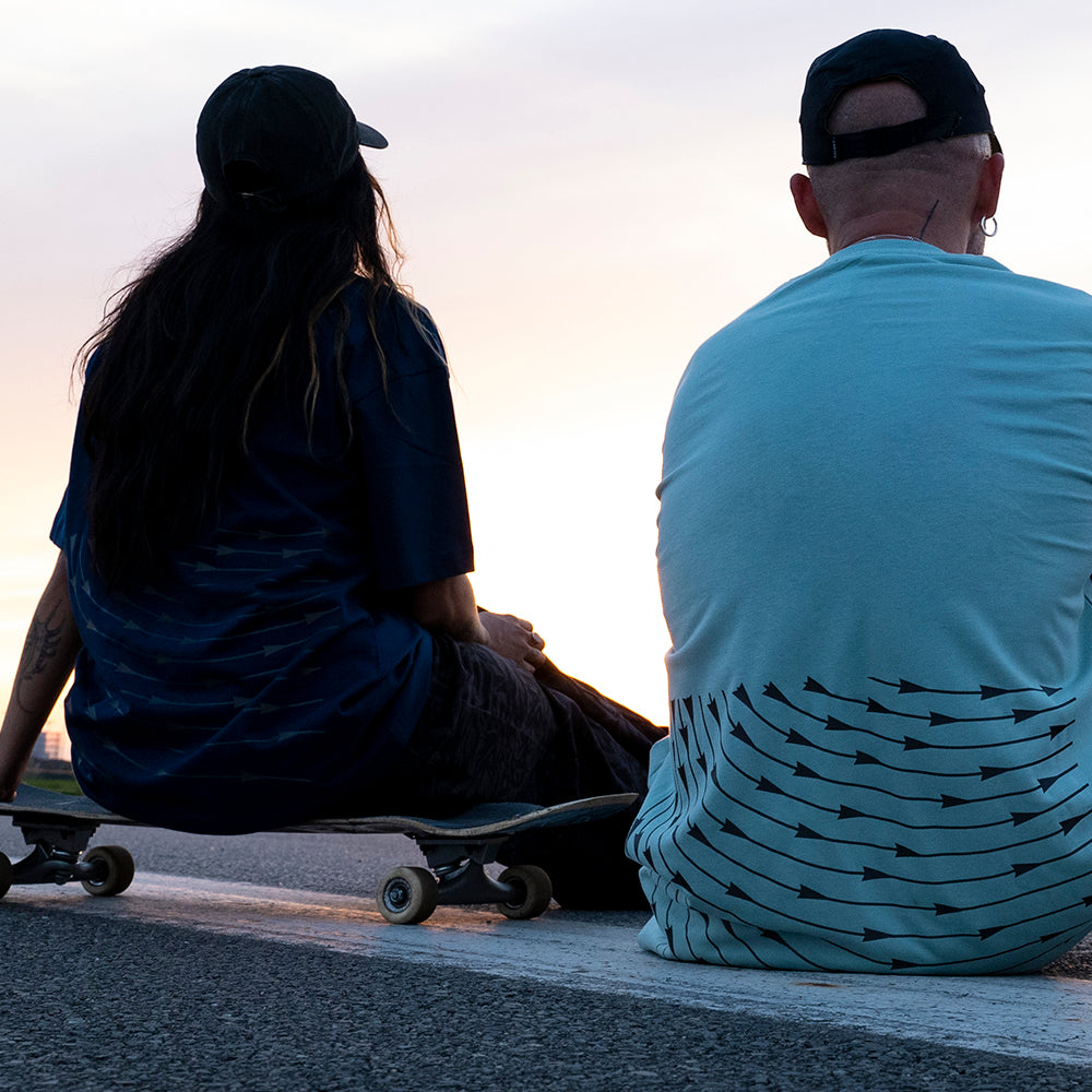 links Frau sitzt auf Skateboard, rechts Mann sitzt auf Boden, Rückenansicht
