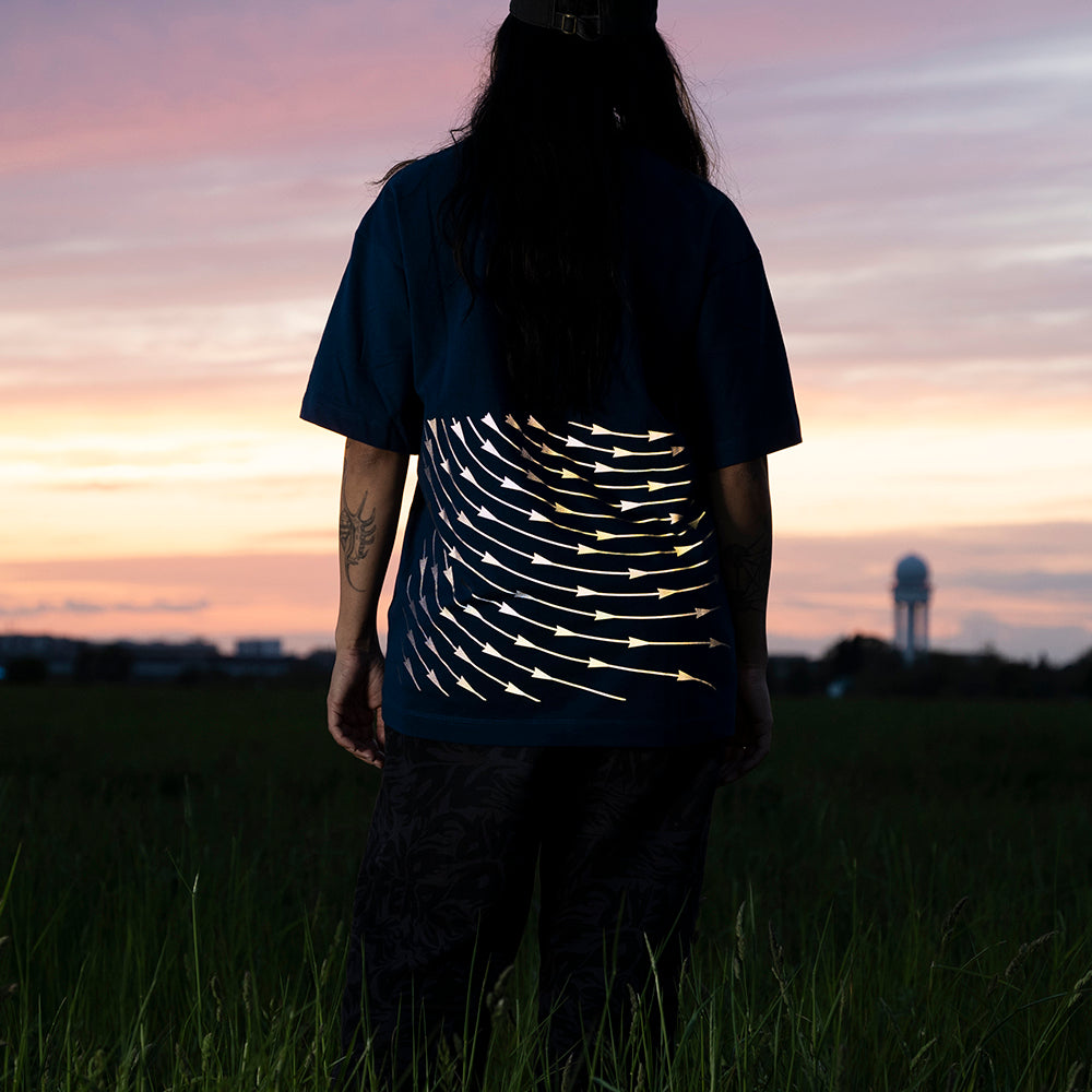 Frau steht im Gras, guckt in den Sonnentuntergang , ihr T-Shirt hat reflektierende Linien