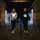 2 sportliche Mädchen gehen unter der U-Bahn entlang, eine mit Fahrrad, eine mit Trinkflasche, beide tragen Kleidung mit reflektierendem Brustprint 