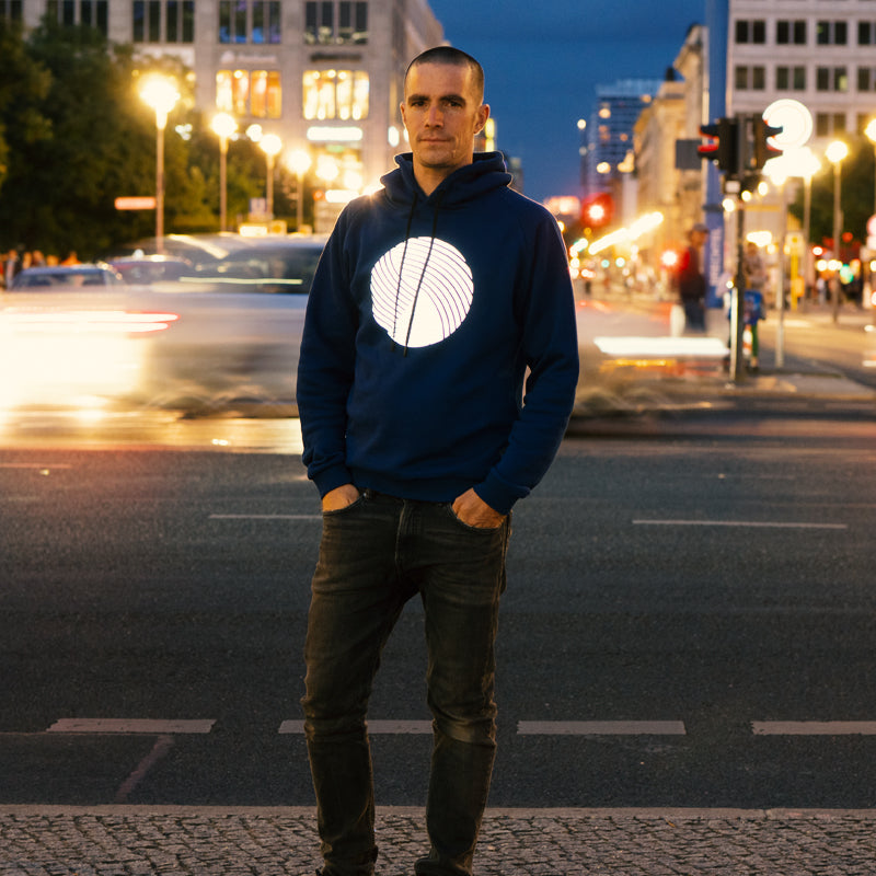 Typ steht vor erleuchteter Straße in der Dämmerung am Potsdamer Platz, trägt dunkelblauen Pullover mit reflektierendem Brustprint