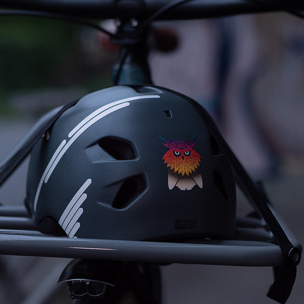 grauer Fahrradhelm auf Frontrack gespannt, versehen mit reflektierenden Stickern, Eule und Streifen